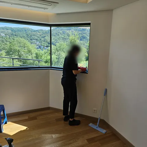 코너 창 앞에서 창틀을 닦는 작업자와 햇살이 드는 원목 마루 방